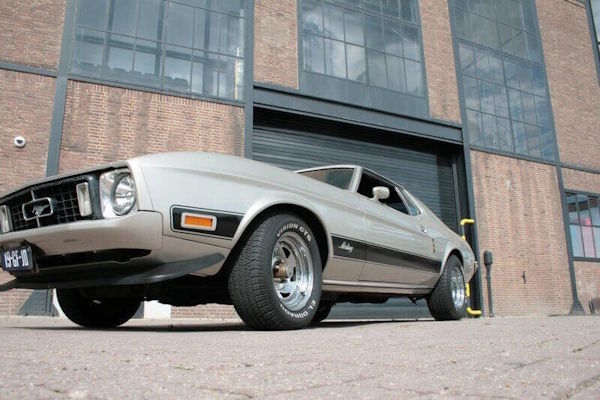 Een statige foto van de hele zilveren Ford Mustang Grande vanuit kikkerperspectief, poserend voor het industriële en monumentale Energiehuis in Dordrecht.