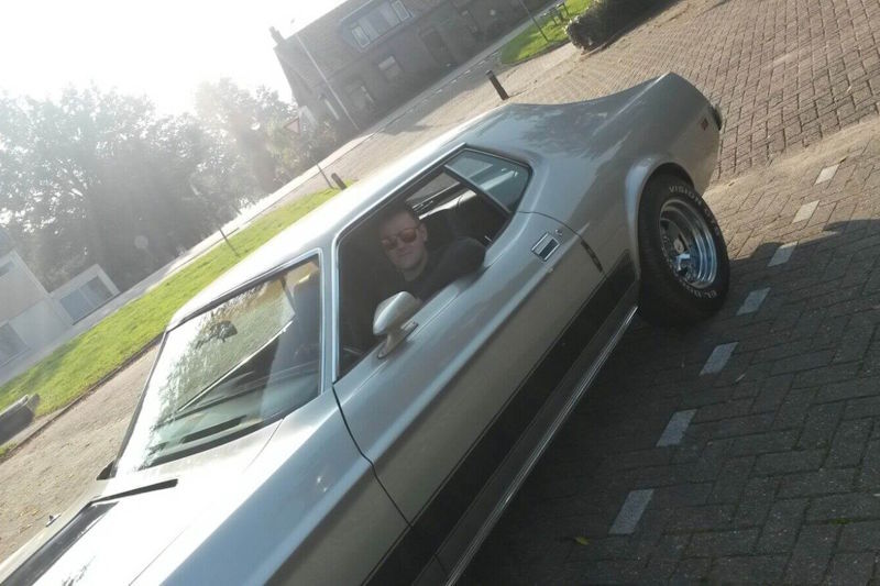 Een trotse man met een glimlach bestuurt de zilveren Ford Mustang Grande en geniet zichtbaar van de V8-roffel en de aandacht tijdens zijn jubileumdag.